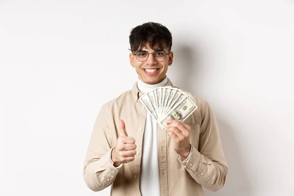 Młody facet w okularach pokazuje dolary i kciuki do góry, zarabiając pieniądze, stojąc z gotówką na białym tle — Zdjęcie stockowe