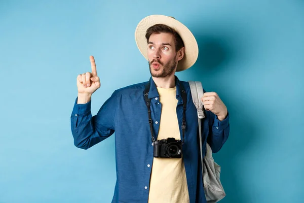 Bonito turista olhando curioso e apontando para cima, mostrando passeios interessantes em férias de verão, vestindo chapéu de palha e segurando mochila com câmera, fundo azul — Fotografia de Stock