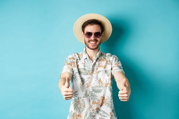 Turista feliz en gafas de sol y sombrero de paja mostrando los pulgares hacia arriba y sonriendo, disfrutando de las vacaciones de verano, de pie sobre fondo blanco — Foto de Stock