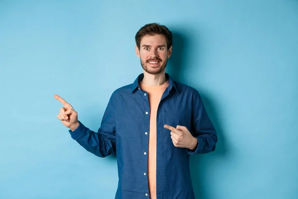 Feliz hombre caucásico sonriente mostrando publicidad, señalando los dedos a la izquierda en el espacio vacío, de pie sobre fondo azul — Foto de Stock