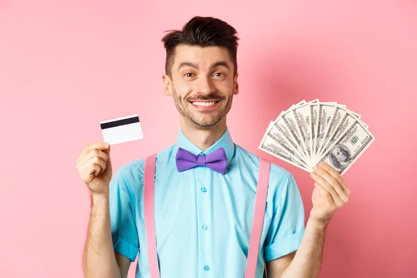 Hombre alegre mostrando su dinero y tarjeta de crédito de plástico, sonriendo feliz a la cámara, de pie sobre fondo rosa — Foto de Stock