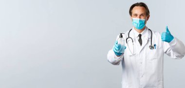 Covid-19, virüsü önleme, sağlık çalışanları ve aşı konsepti. Tıbbi maskeli tatmin olmuş bir doktor ve onaylı eldivenler, el dezenfektanı, beyaz arka plan.