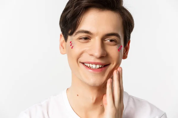 People, lgbtq and beauty concept. Headshot of beautiful gay man with glitter on face, smiling and looking happy, touching cheek after kiss, white background — Stock Photo, Image