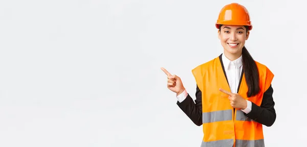 Construcción, construcción y concepto industrial. Sonriente asiática arquitecta en casco de seguridad, ropa reflectante apuntando dedo esquina superior izquierda, mostrando proyecto en el lugar de trabajo, fondo blanco — Foto de Stock