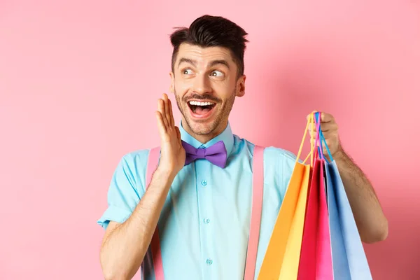 Sorprendido hombre feliz gritando de alegría, mirando a un lado el logotipo y sosteniendo bolsas de compras, de pie sobre el fondo rosa —  Fotos de Stock