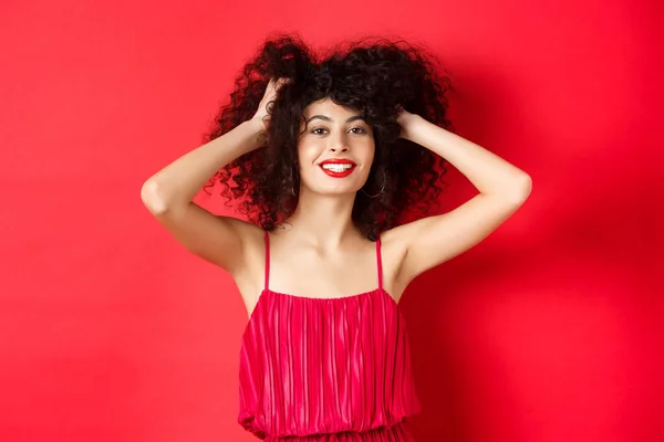 Donna spensierata in abito rosso, toccando i capelli ricci e sorridendo felice, in piedi su sfondo studio — Foto Stock