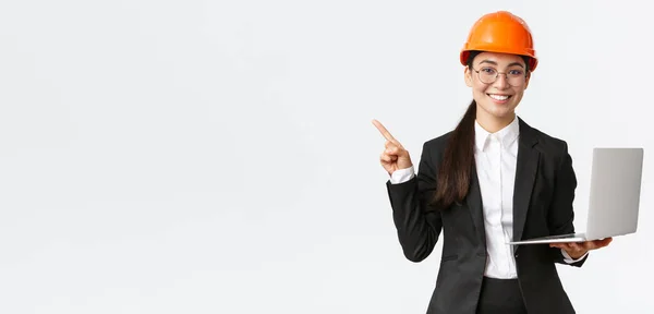 Usmívající se profesionální asijský ženský inženýr nebo architekt ve stavebnictví, nošení bezpečnostní přilby a skafandru, ukazující prstem vlevo při používání notebooku, stojící bílé pozadí — Stock fotografie
