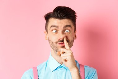 Image of funny caucasian guy picking nose and squint eyes, making dumb face, standing on pink background