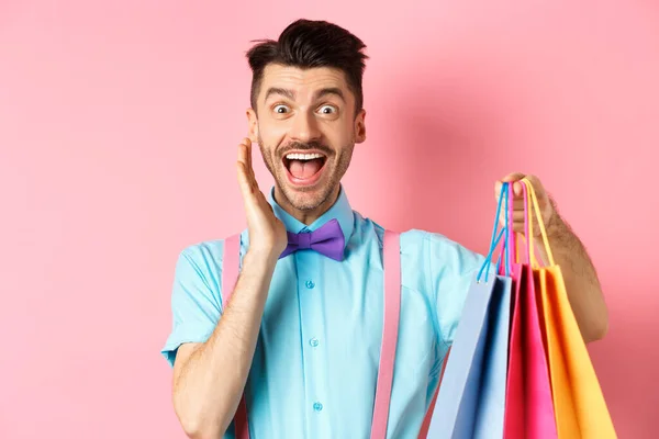 Sorprendido y feliz hombre gritando de alegría y sosteniendo bolsas de compras, comprador ver descuentos, de pie sobre fondo rosa —  Fotos de Stock