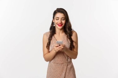 Noel partisi ve kutlama konsepti. Cazibeli güzel bir kadın telefonda mesaj okuyor ve gülümsüyor, akıllı telefon kullanıyor, beyaz arka planda duruyor.