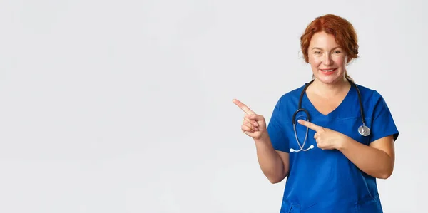 Medycyna, opieka zdrowotna i koncepcja koronawirusa. Portret miłej lekarki, rudowłosa pielęgniarka w niebieskim fartuchu wyglądająca optymistycznie, pokazująca baner kliniki, reklama szpitala, wskazująca palcem w lewo — Zdjęcie stockowe