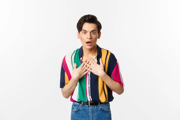 Portrait of surprised gay man holding hands on heart, looking with disbelief and amazement at camera, standing over white background — Stock Photo, Image