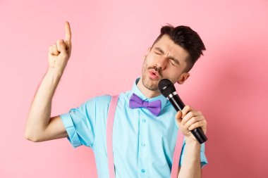 Parti ve festival konsepti. Komik adam mikrofonda şarkı söylüyor, parmağını kaldırıyor şarkıda yüksek notaya ulaşıyor, pembe arka planda duruyor.