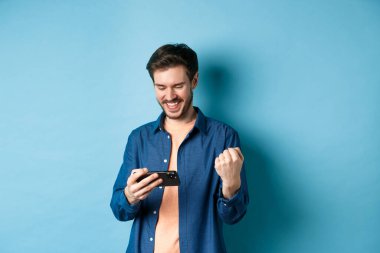 Cep telefonuyla video oyununda kazanan mutlu genç adam, akıllı telefon ekranına bakıyor ve sevinçle evet diyor, mavi arka planda duruyor.