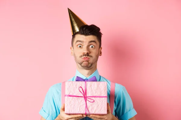 Vacaciones y concepto de celebración. Chico divertido mirando a la cámara sorprendida, usando sombrero de fiesta, sosteniendo regalo de cumpleaños y conteniendo la respiración, haciendo pucheros en la cámara, fondo rosa — Foto de Stock