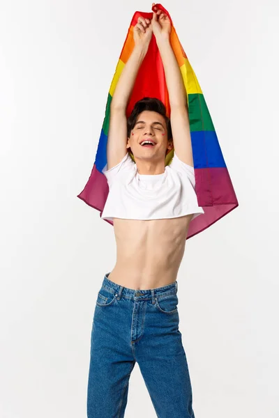 Vista vertical del hombre gay orgulloso y feliz levantando la bandera del arco iris lgbtq, sonriendo con emoción aliviada, usando top con jeans, fondo blanco — Foto de Stock