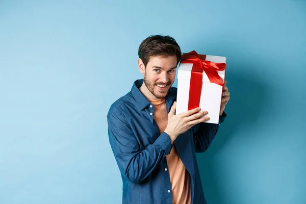 День Святого Валентина. Романтический парень показывает подарочную коробку для любовника, празднование годовщины, стоя на синем фоне — стоковое фото