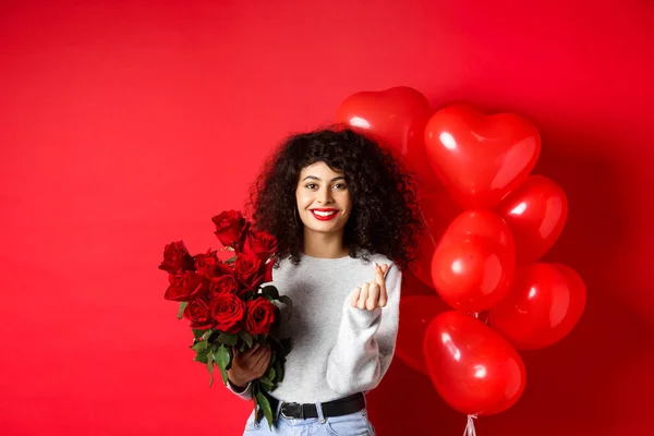 Holidays and celebration. Beautiful girlfriend receive flowers on anniversary, showing finger heart and standing near party balloons, red background