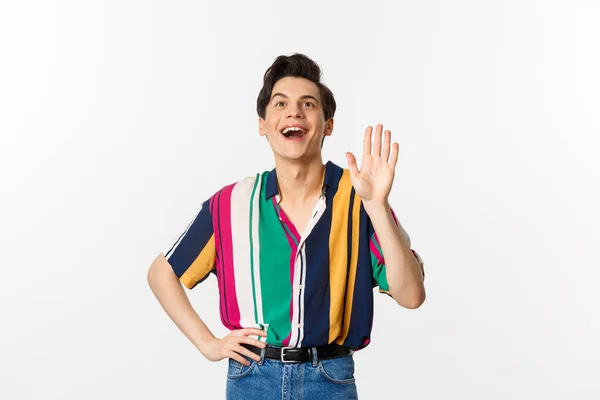 Cheerful gay man saying hello, waving hand and greeting person, standing over white background — Stock Photo, Image