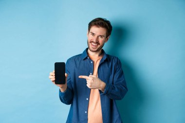 Boş akıllı telefon ekranında gülümseyen beyaz adam, cep telefonunu işaret ediyor ve memnun görünüyor, mavi arka planda duruyor.
