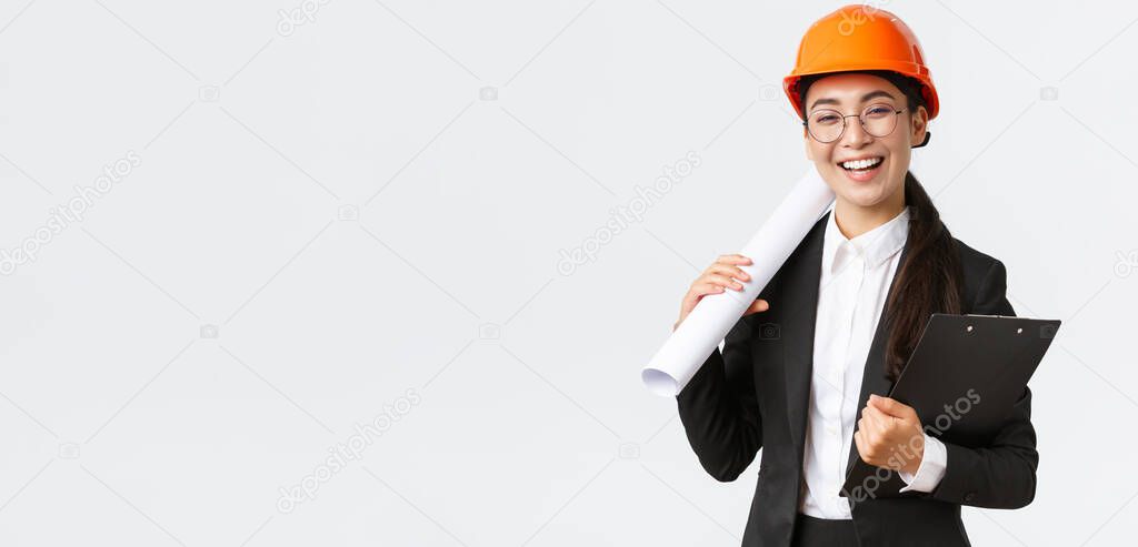 Professional happy asian female architect, construction engineer in helmet and business suit holding blueprints and clipboard with building documentation, smiling cheerful, white background