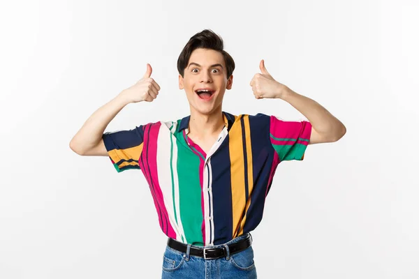 Excited and amazed young man showing thumbs up with impressed expression, standing over white background — Stock Photo, Image