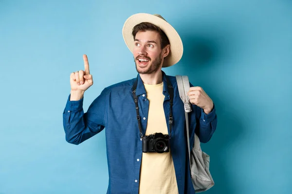 Bello turista che guarda e punta in alto, viaggia in vacanza estiva con zaino e macchina fotografica, in piedi su sfondo blu — Foto Stock