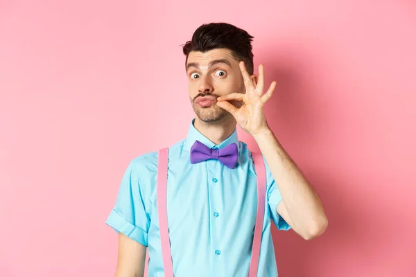Смішний молодий чоловік торкається французьких вусів і почервонілих губ, дивлячись тупо на камеру, стоїть в лапці і підтяжки на рожевому фоні — стокове фото