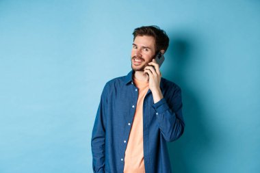 Modern gülümseyen adam cep telefonuyla konuşuyor, kameraya mutlu görünüyor, mavi arka planda duruyor.