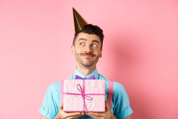Vacaciones y concepto de celebración. Chico tonto con bigote y pajarita, usando sombrero de fiesta, recibir regalo de cumpleaños y mirando soñador a un lado, de pie sobre fondo rosa — Foto de Stock