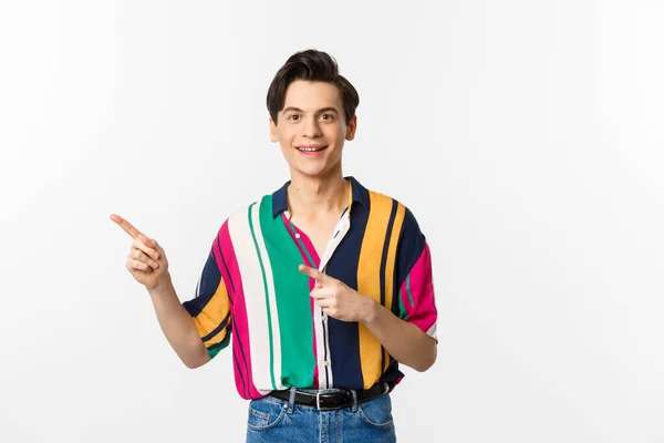 Arckép egy jóképű fiatalember elegáns ruhákban, ujjal mutogatva balra és mosolyogva, hirdetést mutat, fehér háttér felett áll — Stock Fotó