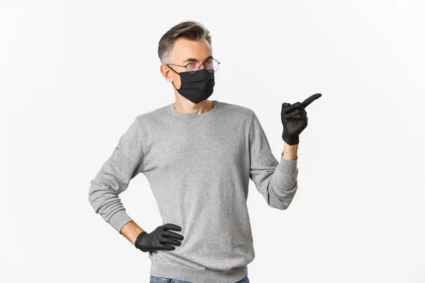 Concept de coronavirus, mode de vie et quarantaine. Homme d'âge moyen intrigué en masque médical, gants et lunettes pointant du doigt à droite et regardant la publicité, debout sur fond blanc — Photo