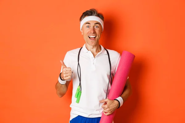 Konzept aus Workout, Fitness und Lifestyle. Fröhlicher Fitness-Typ mittleren Alters, der Springseil und Yogamatte in der Hand hält, Daumen nach oben zeigt und zufrieden lächelt, vor orangefarbenem Hintergrund steht — Stockfoto