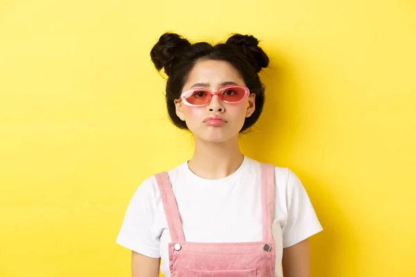 Léto a módní koncept. znuděný asijské dospívající dívka v sluneční brýle trucování, stojící náladový na žlutém pozadí — Stock fotografie