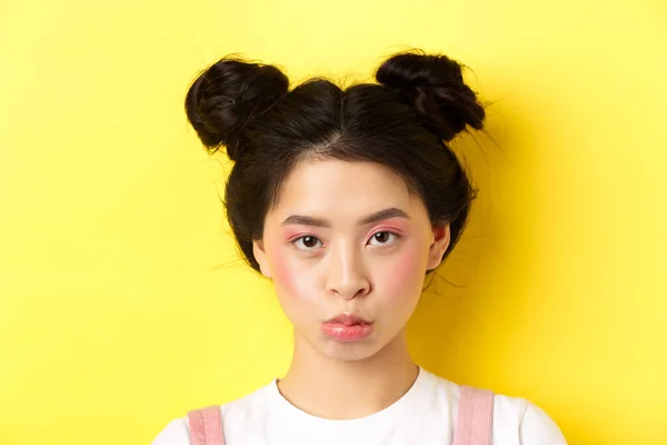 Close up ritratto di lunatico asiatico ragazza pouting e looking sconvolto a camera, standing wiht glamour trucco e hairbuns su fondo giallo — Foto Stock