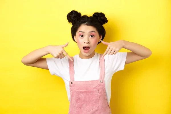 Excited teenage asian girl with beauty makeup, pointing fingers down and open mouth fascinated, standing happy on yellow background — Stock Photo, Image