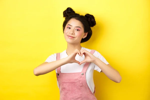 Ημέρα του Αγίου Βαλεντίνου. Χαριτωμένο κορίτσι από την Ασία στέλνει την αγάπη της, δείχνει χειρονομία καρδιάς και χαμογελά στην κάμερα ρομαντική, στέκεται σε κίτρινο φόντο — Φωτογραφία Αρχείου