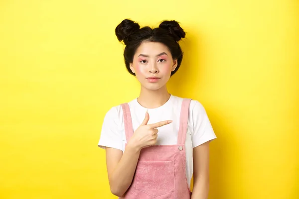 Elegante ragazza adolescente asiatica con trucco e vestiti estivi, indicando il dito destro e guardare serio, in piedi contro lo sfondo giallo — Foto Stock