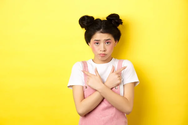 Indeciso timido asiatico ragazza indicando lateralmente, mostrando sinistra e destra modi e broncio sconvolto, in piedi triste su sfondo giallo — Foto Stock
