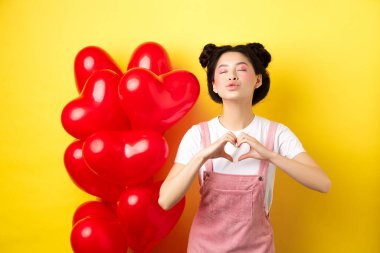Romantik kız, gözlerini kapat, dudaklarını büzüştür, seni sevdiğimi göster, kalp jestini göster, sevimli kırmızı balonların yanında dur, sarı arka planda sevgiliyi öp.