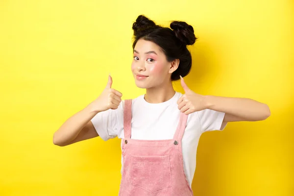 Hermosa chica asiática con maquillaje rosa brillante, mostrando el pulgar hacia arriba en la aprobación y sonriendo satisfecho, recomendando promo, fondo amarillo —  Fotos de Stock
