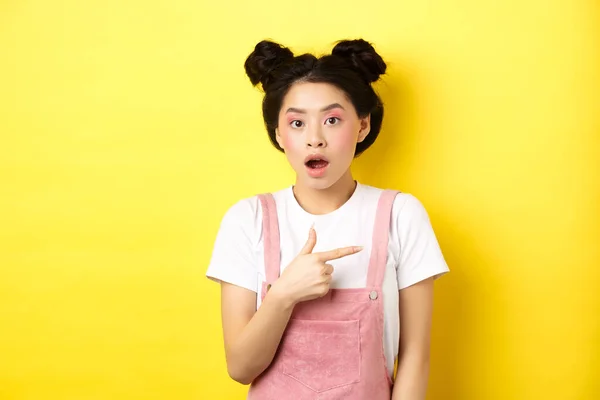 Scioccato donna asiatica con trucco glamour, bocca aperta e puntando a destra al logo, mostrando pubblicità, in piedi su sfondo giallo — Foto Stock