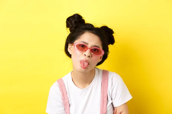Καλοκαίρι και ομορφιά έννοια. Παιδική Ασιάτισσα με γυαλιά ηλίου που δείχνει γλώσσα, είναι αγενής, στέκεται σε κίτρινο φόντο — Φωτογραφία Αρχείου