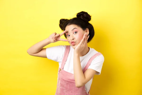 Hermosa chica asiática mostrando v-signo y haciendo pucheros lindo, haciendo cara tonta con maquillaje, de pie sobre fondo amarillo —  Fotos de Stock