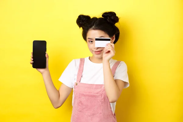 Online shopping concept. Cute asian girl paying with plastic credit card, showing empty smartphone screen and smiling, yellow background — Stock Photo, Image