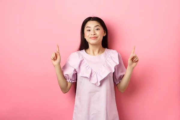 Hermosa joven mujer asiática en vestido romántico señalando los dedos hacia arriba, sonriendo y mostrando con gusto anuncio, de pie sobre fondo rosa — Foto de Stock