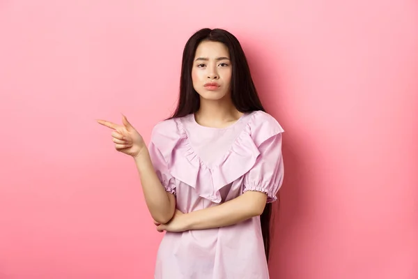 Σκεπτικό κορίτσι από την Κίνα δείχνει το δάχτυλο αριστερά, δείχνει αδιάφορη και σοβαρή, δείχνει την προσφορά, στέκεται στο φόρεμα σε ροζ φόντο — Φωτογραφία Αρχείου