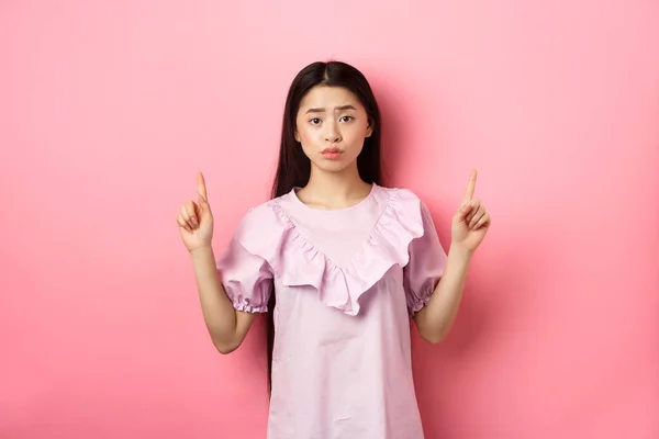 Mujer asiática tímida lindo señalando los dedos hacia arriba, frunciendo el ceño y mirar molesto, señalando los dedos hacia arriba en el logotipo, de pie en vestido sobre fondo rosa —  Fotos de Stock
