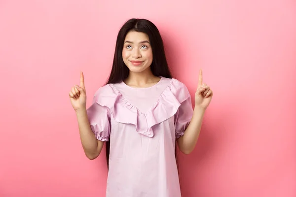 Όμορφη χαμογελαστή Ασιάτισσα έφηβη που δείχνει τα δάχτυλα ψηλά, κοιτάζει διαφήμιση με χαρούμενο πρόσωπο, στέκεται σε ροζ φόντο — Φωτογραφία Αρχείου