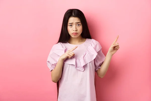 Triste y angustiado chica asiática frunciendo el ceño, enfurruñando y señalando los dedos a la derecha en decepcionantes malas noticias, sintiéndose infeliz, de pie sobre el fondo rosa —  Fotos de Stock